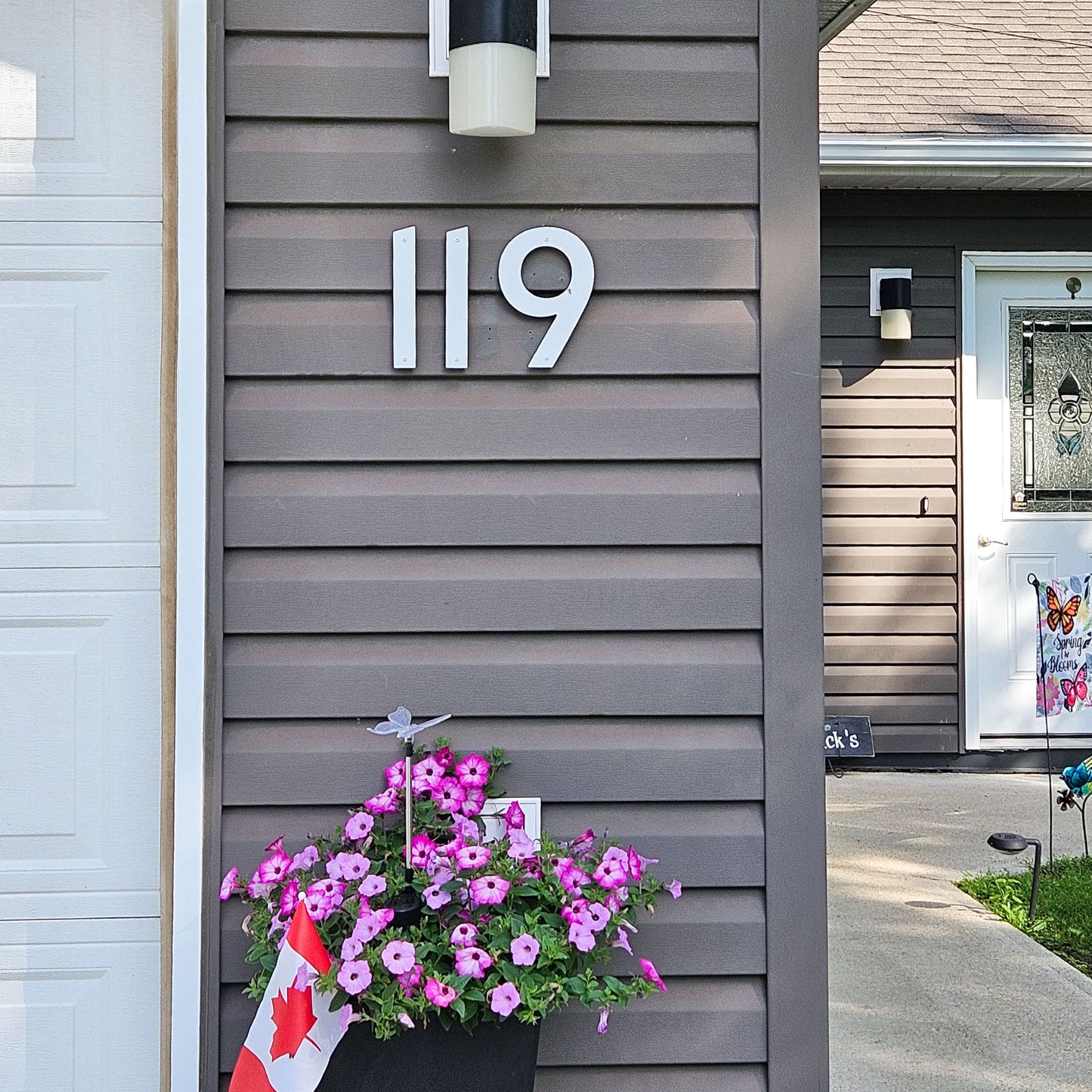 8 inch CLASSIC MODERN White house numbers '119' mounted on a dark gray horizontal siding wall, with a flower box of pink petunias and a small Canadian flag at the bottom left.