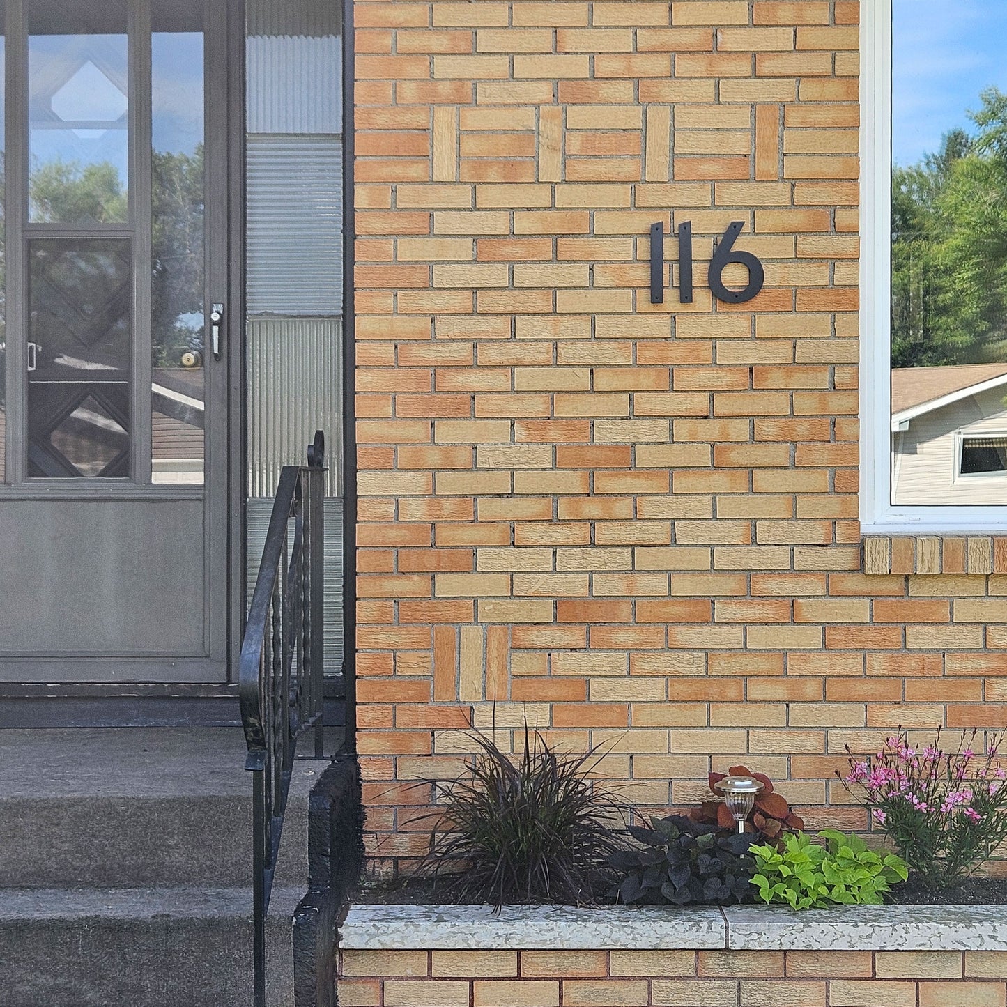 A brick house exterior with the black CLASSIC MODERN house numberS 116 mounted on a tan brick wall. A glass front door with a black metal railing is on the left, and a small garden bed with plants and a small decorative figure is at the bottom right.
