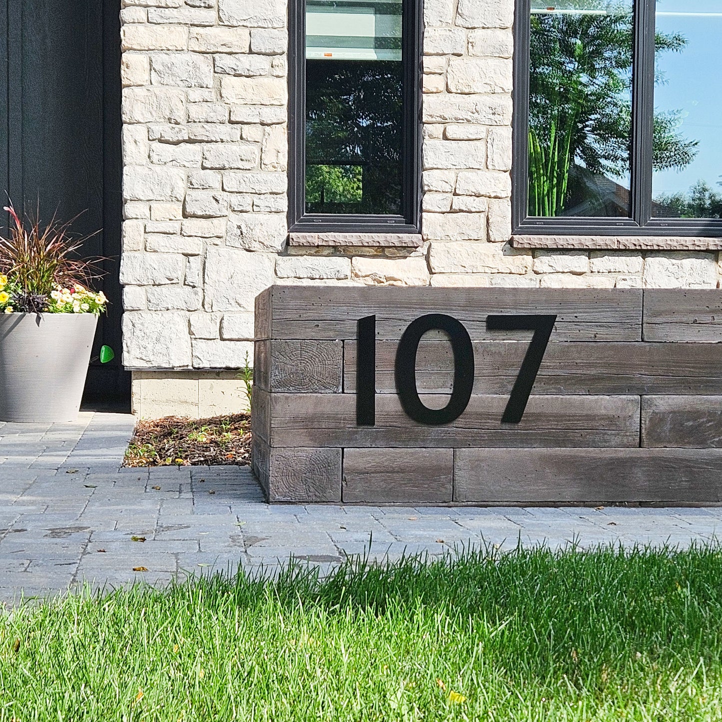 Large 12 inch CLASSIC MODERN black plastic house numbers '107' mounted on a rustic gray wooden retaining wall in front of a light stone house exterior.