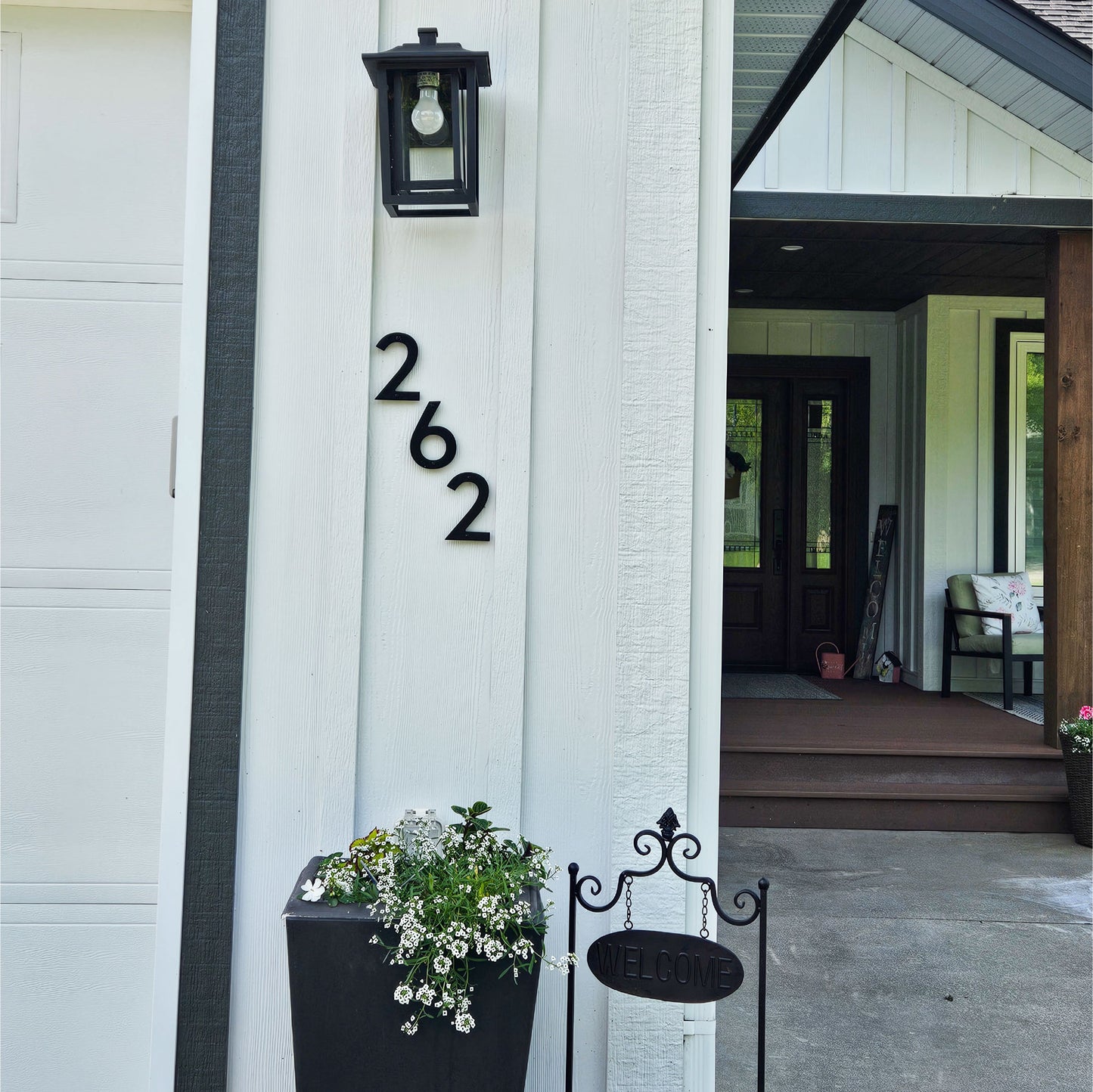 5 inch CLASSIC MODRN Black house numbers '262' mounted on a white exterior wall next to a black lantern light fixture and a potted plant, with a front porch visible in the background.