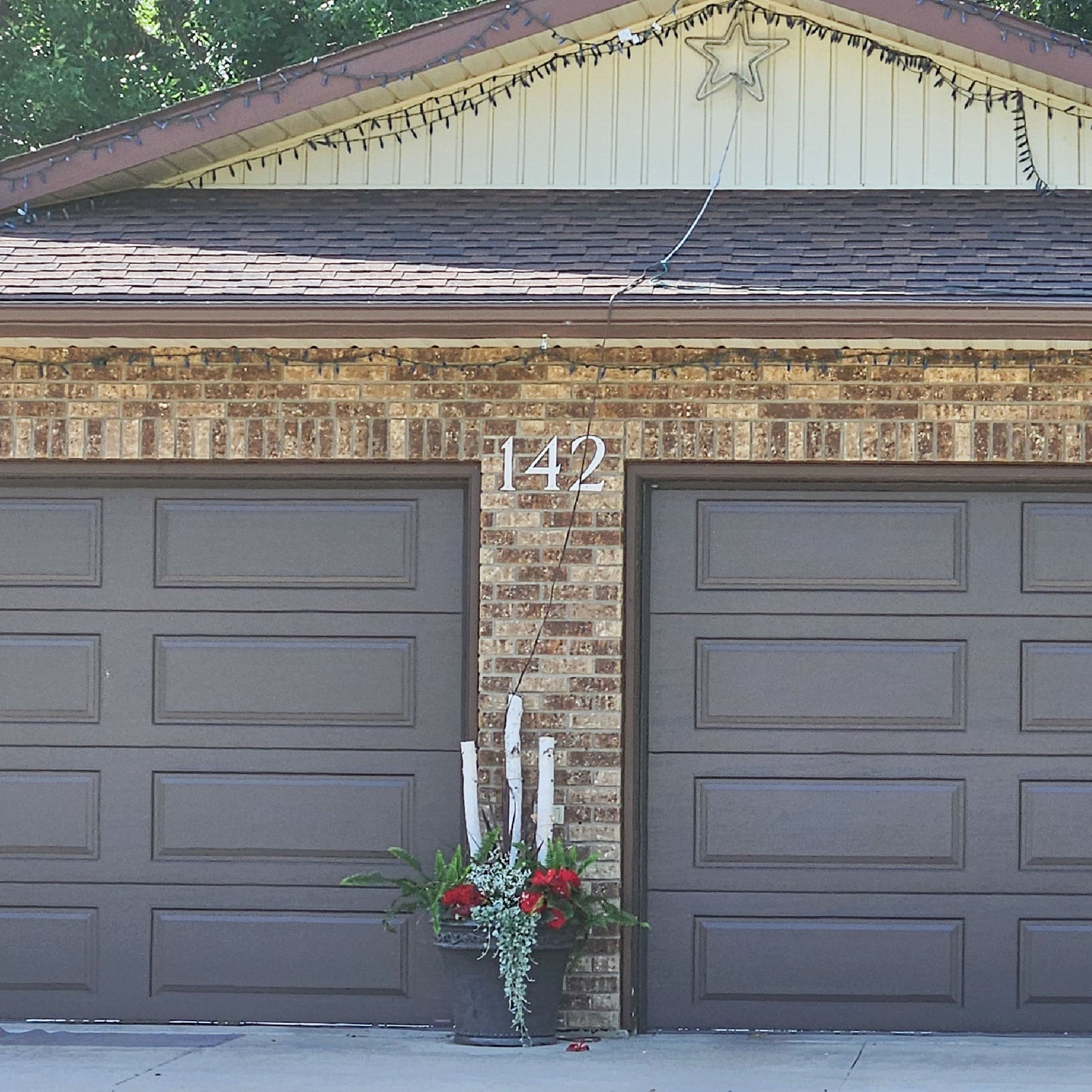 A front-facing view of a two-car garage with dark brown paneled doors,  the silver ROMAN SERIF house number 142 between the two garage doors on the brick siding