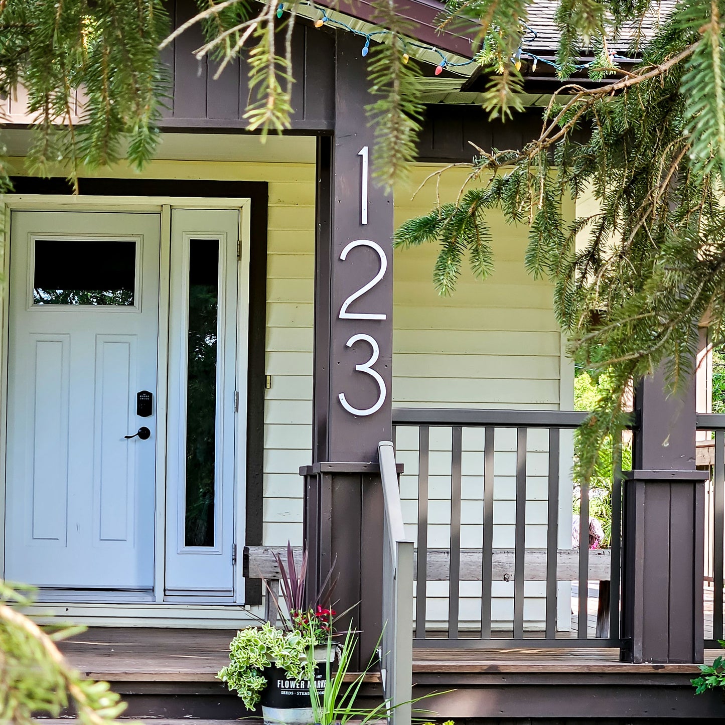 A front porch entryway with a white door, pale yellow siding, dark brown trim and posts, a 12 inch tall white house numbers  123 fastened vertically on the post, and evergreen branches hanging over the scene.