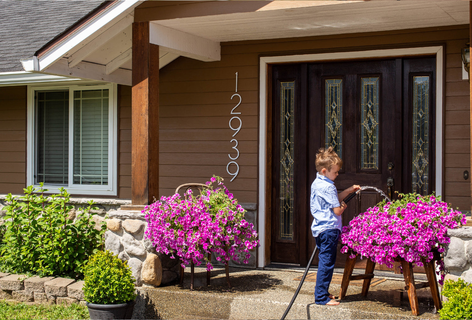 THIN MODERN white house numbers on brown siding while a boy waters pink petunias